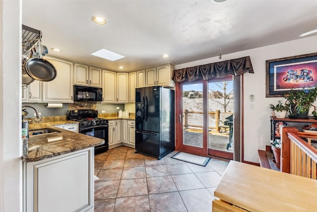 kitchen with dark stone counters, black appliances, sink, a skylight, and cream cabinetry