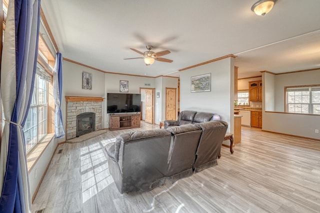 living area with light wood finished floors, baseboards, a ceiling fan, crown molding, and a stone fireplace