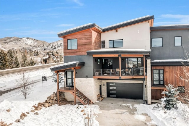 snow covered back of property featuring a mountain view, stucco siding, an attached garage, and driveway