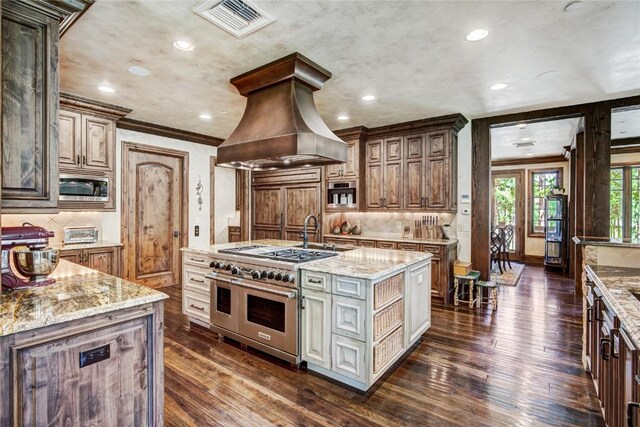 kitchen featuring stainless steel appliances, light stone counters, dark hardwood / wood-style flooring, premium range hood, and a kitchen island with sink