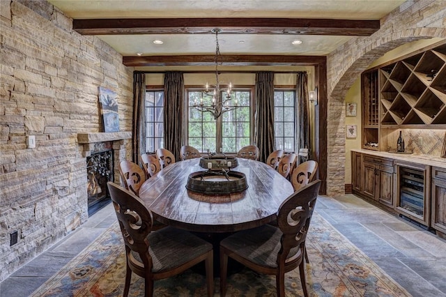 dining space with beamed ceiling, a notable chandelier, a stone fireplace, and beverage cooler