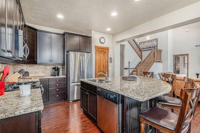 kitchen with light stone counters, appliances with stainless steel finishes, dark hardwood / wood-style flooring, and an island with sink