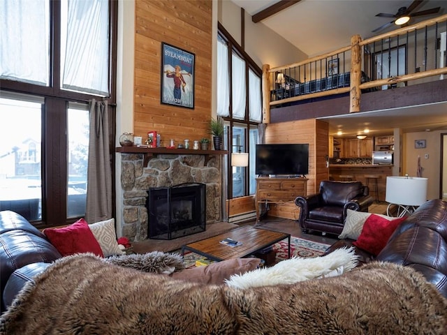 living room with a high ceiling, wood walls, beam ceiling, ceiling fan, and a stone fireplace