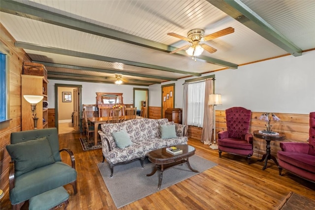 living area with wooden walls, beamed ceiling, wainscoting, wood finished floors, and a ceiling fan