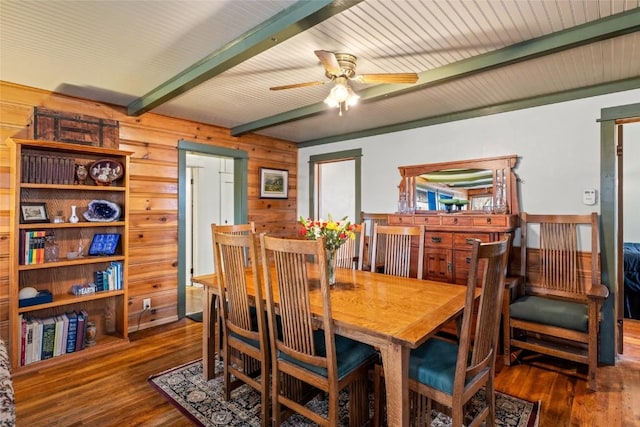 dining room featuring wooden walls, beamed ceiling, a ceiling fan, and wood finished floors