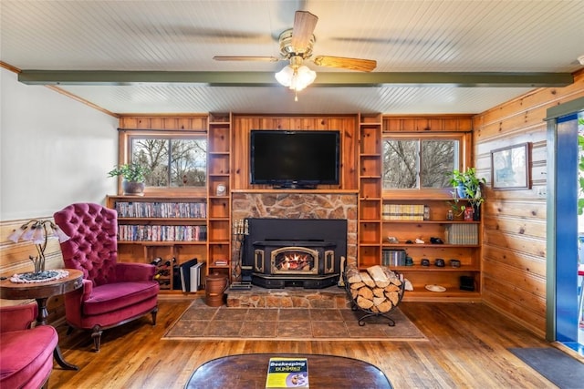 living area with ceiling fan, wood walls, beam ceiling, a wood stove, and hardwood / wood-style flooring