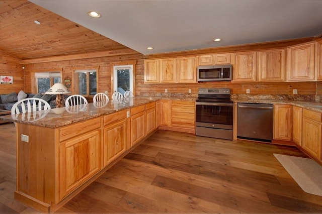 kitchen with wood walls, appliances with stainless steel finishes, kitchen peninsula, light stone countertops, and light hardwood / wood-style floors