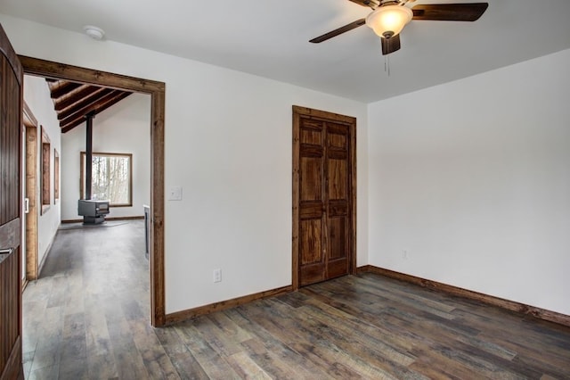 empty room featuring baseboards, a ceiling fan, lofted ceiling with beams, wood finished floors, and a wood stove