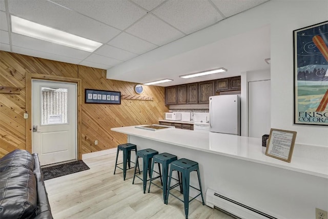 kitchen featuring a paneled ceiling, white appliances, a baseboard radiator, a kitchen bar, and kitchen peninsula