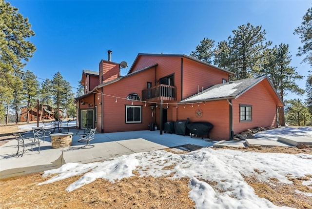 snow covered rear of property with a balcony, a patio area, a chimney, and a fire pit