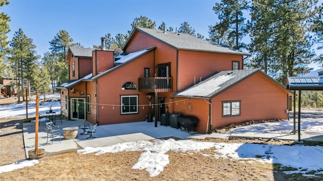 snow covered back of property with a fire pit, a patio, a balcony, a chimney, and roof with shingles