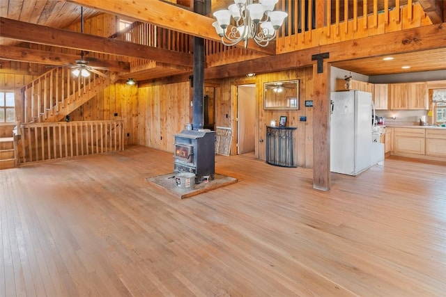 unfurnished living room with ceiling fan with notable chandelier, light wood-type flooring, a wood stove, and wooden walls