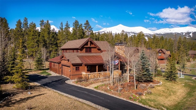 chalet / cabin featuring a wooded view, a porch, a chimney, aphalt driveway, and a mountain view