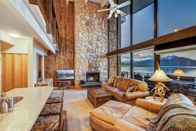 living room featuring carpet flooring, a towering ceiling, ceiling fan, wooden walls, and a stone fireplace