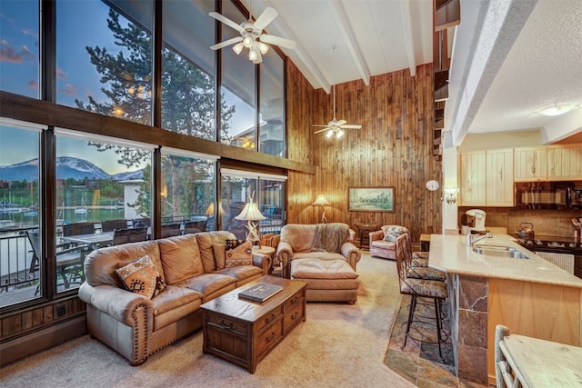 carpeted living room featuring wood walls, high vaulted ceiling, sink, ceiling fan, and beamed ceiling