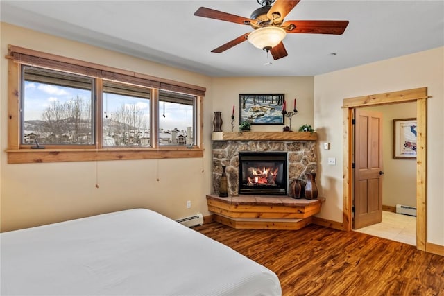 bedroom with a stone fireplace, baseboard heating, light wood-type flooring, and ceiling fan