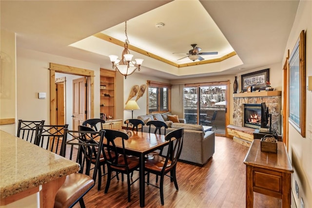 dining area with ceiling fan with notable chandelier, a stone fireplace, wood-type flooring, and a tray ceiling