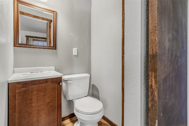 half bath featuring toilet, a textured wall, baseboards, and vanity