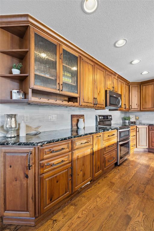 kitchen featuring dark wood-style flooring, appliances with stainless steel finishes, brown cabinetry, dark stone countertops, and glass insert cabinets