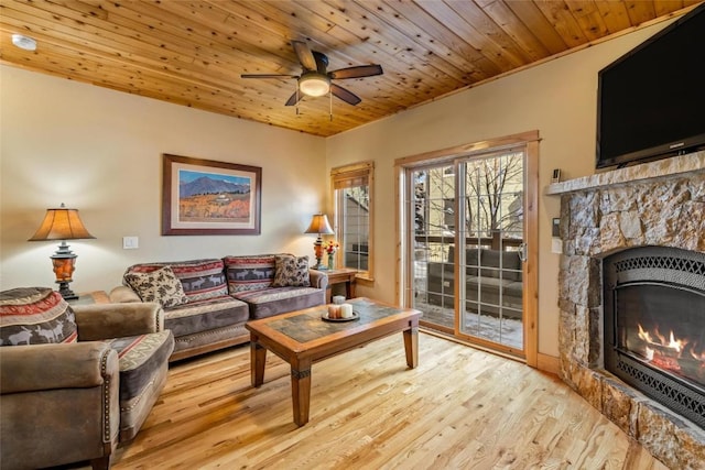 living area featuring ceiling fan, light wood-type flooring, a stone fireplace, and wooden ceiling