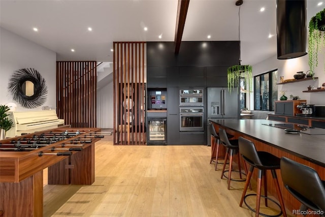 kitchen with appliances with stainless steel finishes, a kitchen bar, beamed ceiling, wine cooler, and light hardwood / wood-style floors