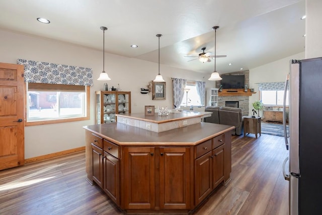 kitchen with stainless steel refrigerator, ceiling fan, a center island, a brick fireplace, and dark hardwood / wood-style flooring