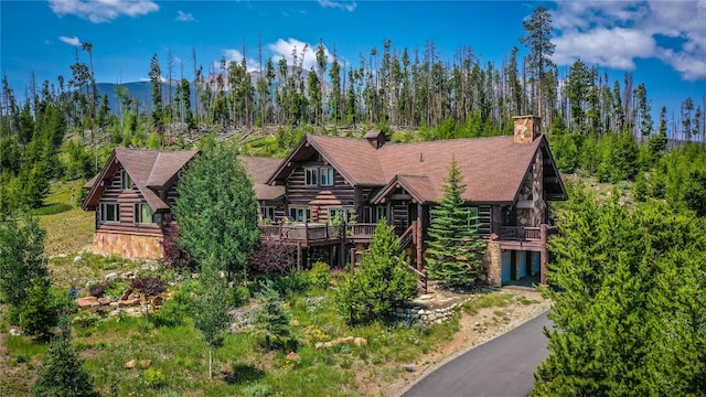 cabin featuring a deck, driveway, log siding, a forest view, and a chimney