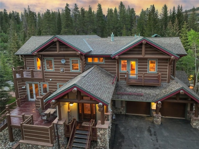 view of front of house with a balcony, a forest view, driveway, an attached garage, and a shingled roof