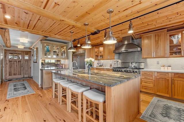 kitchen with wall chimney range hood, stainless steel built in fridge, dark stone countertops, backsplash, and a center island with sink