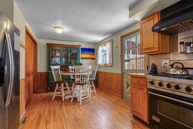 kitchen with wall chimney exhaust hood, light wood-type flooring, stainless steel appliances, and wooden walls