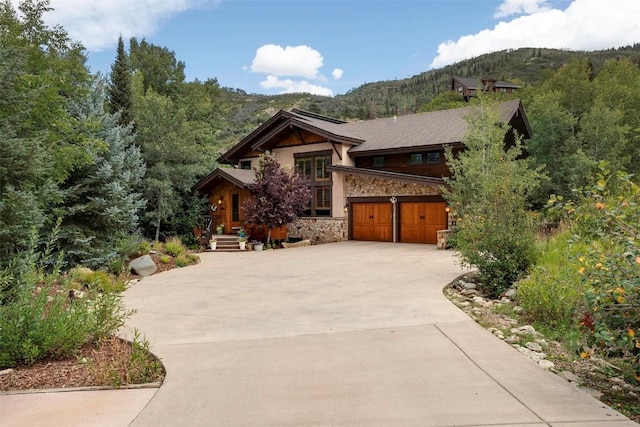 view of front of house with a garage, stone siding, driveway, and a wooded view