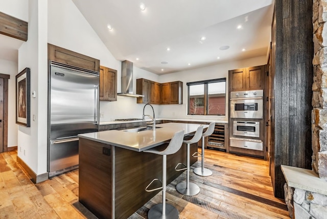 kitchen featuring stainless steel appliances, a kitchen island with sink, sink, wall chimney range hood, and a breakfast bar area