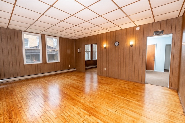 empty room featuring a paneled ceiling, light wood-type flooring, baseboard heating, and wood walls