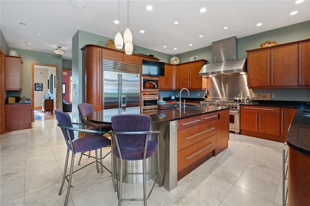 kitchen with an island with sink, wall chimney exhaust hood, built in appliances, light tile patterned floors, and pendant lighting