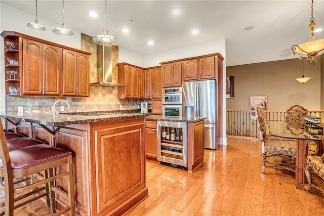 kitchen featuring hanging light fixtures, stainless steel appliances, wine cooler, wall chimney exhaust hood, and light wood-type flooring