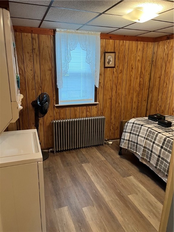 bedroom with a drop ceiling, wood walls, radiator, stacked washer / dryer, and dark hardwood / wood-style floors