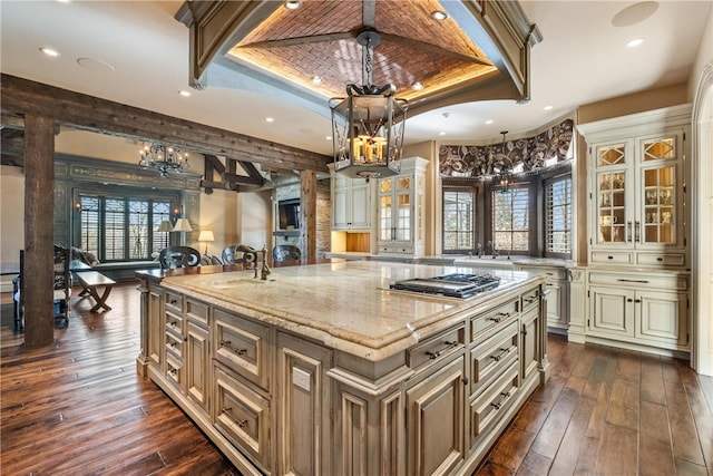 kitchen with a tray ceiling, light stone countertops, a center island with sink, and dark wood-type flooring