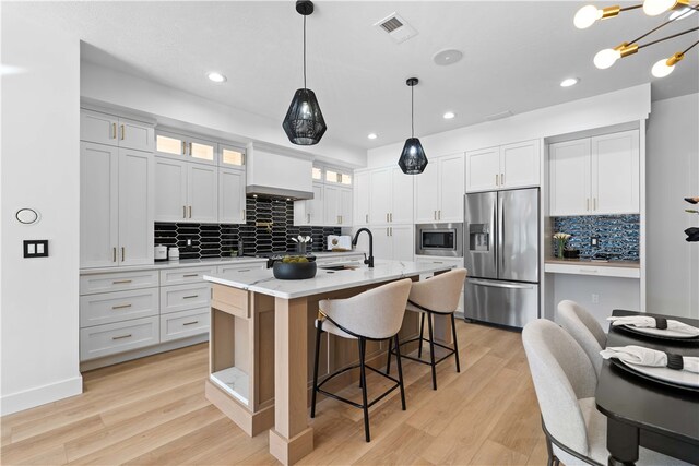 kitchen featuring light hardwood / wood-style floors, white cabinets, a center island with sink, stainless steel appliances, and pendant lighting