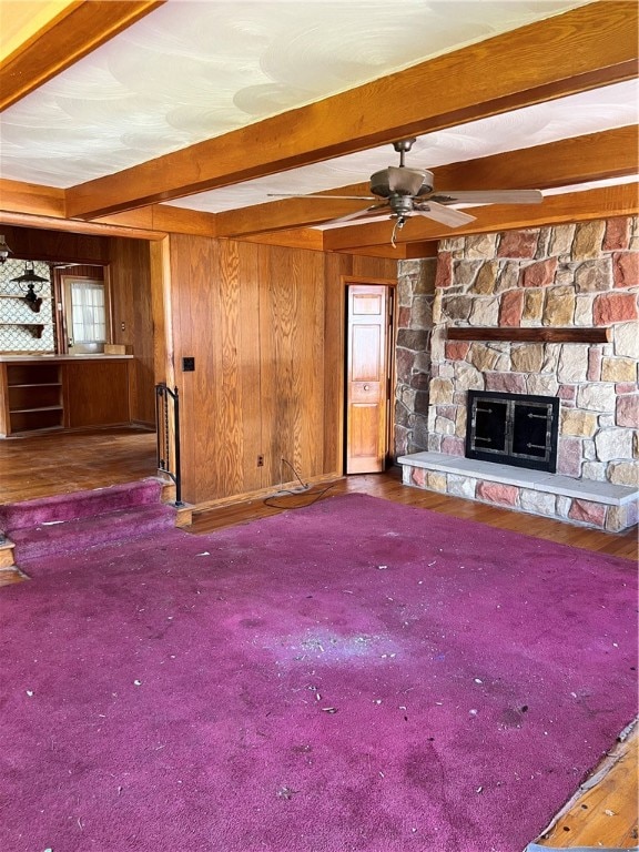 unfurnished living room featuring wood walls, a stone fireplace, dark colored carpet, and beamed ceiling