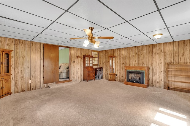 unfurnished living room featuring carpet, wooden walls, ceiling fan, and a drop ceiling