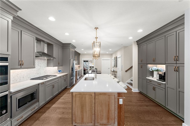 kitchen featuring wall chimney range hood, gray cabinets, dark hardwood / wood-style flooring, stainless steel appliances, and a kitchen island with sink