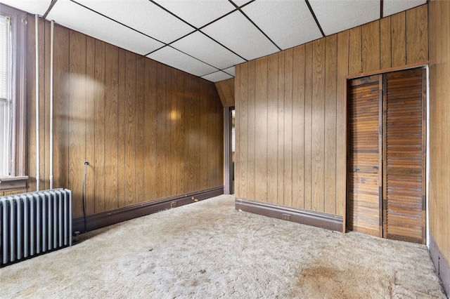 carpeted spare room featuring wood walls, a drop ceiling, and radiator heating unit