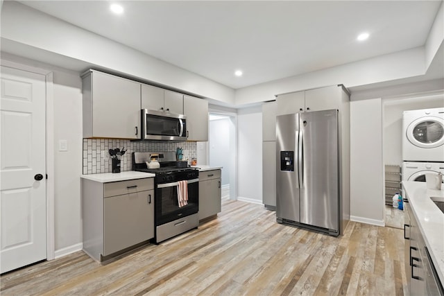 kitchen with gray cabinetry, light hardwood / wood-style flooring, stacked washer / drying machine, stainless steel appliances, and tasteful backsplash