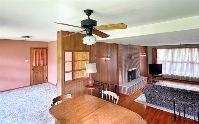 dining room with a brick fireplace, ceiling fan, brick wall, wooden walls, and dark carpet