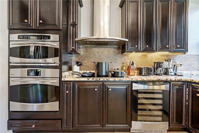 kitchen with wall chimney exhaust hood, stainless steel double oven, tasteful backsplash, and beverage cooler