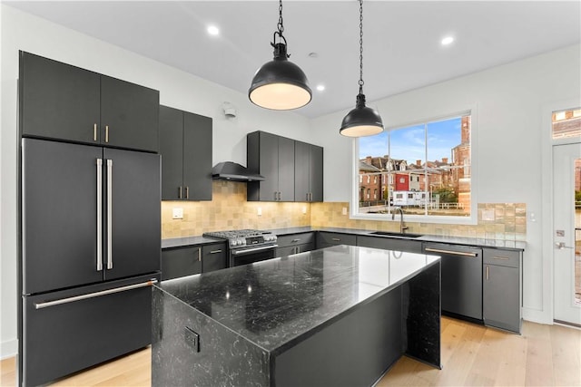 kitchen featuring wall chimney range hood, sink, appliances with stainless steel finishes, a kitchen island, and decorative light fixtures