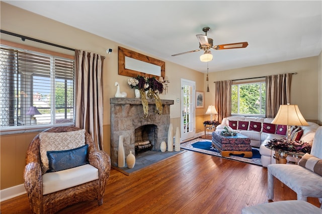 living room with a stone fireplace, dark wood-type flooring, and ceiling fan