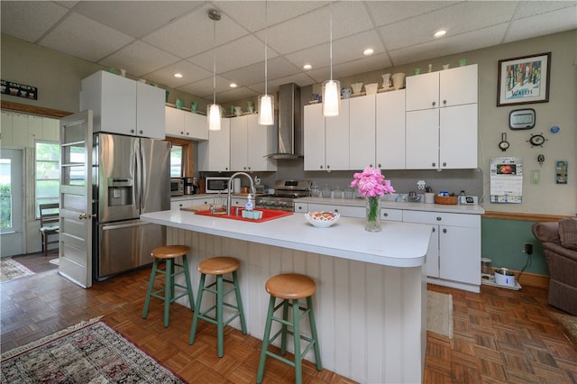 kitchen featuring appliances with stainless steel finishes, a center island with sink, dark parquet flooring, wall chimney range hood, and white cabinetry