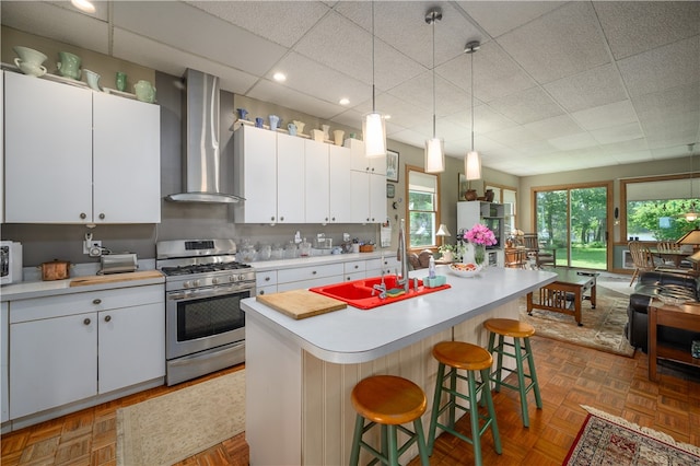 kitchen with wall chimney exhaust hood, gas range, white cabinetry, a center island with sink, and parquet floors