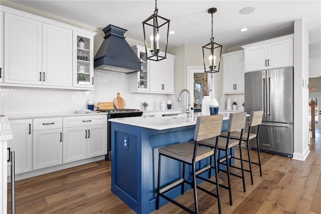 kitchen with custom range hood, hardwood / wood-style flooring, a kitchen island with sink, high end fridge, and backsplash